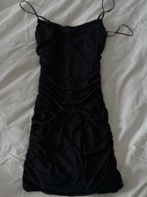 Svart omlottklänning från BikBok - Svart kort klänning från BikBok med tunna axelband och omlottdetalj. Klänningen har en figurnära passform och rynkade partier längs sidorna för en snygg siluett. Perfekt för dig som gillar en enkel men stilren look.