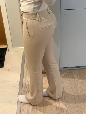 Beige kostymbyxa med utsvängda ben - Snygga beige kostymbyxor med utsvängda ben. Från design by si, fläck på benet men går bort i tvätten. Sitter typ som en XS