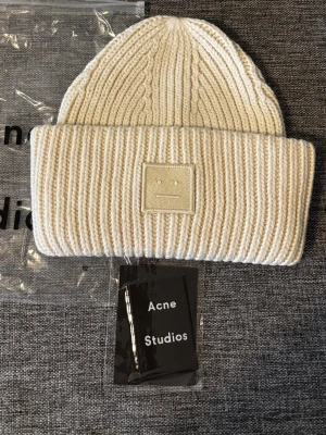 Acne studios mössa - Säljer en vit ribbstickad mössa från Acne Studios med uppvikt kant och det klassiska ansiktslogotypen framtill. Mössan är tillverkad i mjukt material och har en clean, minimalistisk look som passar perfekt till streetwear och vardagsstil.