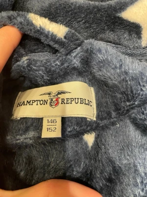 Mjuk blå morgonrock med stjärnor - Supermjuk och gosig blå morgonrock från Hampton Republic med vita stjärnor över hela plagget. Morgonrocken har två stora fickor framtill och ett matchande midjebälte. Perfekt för att mysa runt hemma under kyliga dagar.