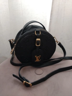 LV monogram svart rund handväska - Säljer en svart rund handväska från LV i präglat läder med guldiga metalldetaljer och justerbar axelrem. Väskan har det klassiska LV-märket framtill och dragkedja upptill.  Fin patina och naturligt slitage. Ingen lukt och kommer från rök och djurfritt hem.