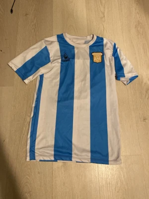 Argentina Maradona matchtröja - Säljer en klassisk Argentina matchtröja från Le Coq Sportif med blå och vita vertikala ränder. På bröstet finns AFA-emblemet och på ryggen står det Maradona samt nummer 10 i svart. Perfekt för fotbollsfans som vill sticka ut på planen eller läktaren.
