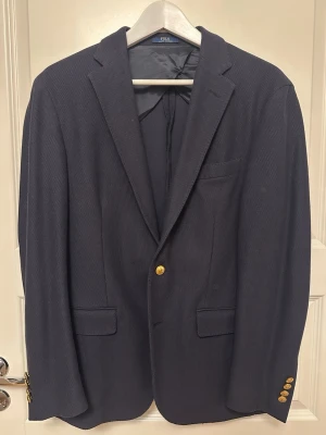 Polo Ralph Lauren Kavaj - Stilren mörkblå kavaj från Polo Ralph Lauren med klassisk passform, guldfärgade knappar och diskret struktur i tyget. Storlek 40R / 50EU