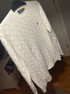 Vit kabelstickad tröja Polo Ralph Lauren - Kabelstickad vit tröja från Polo Ralph Lauren med rund halsringning och den klassiska broderade loggan i marinblått på bröstet. Tröjan har långa ärmar och ribbade muddar vid ärmslut och nederkant. Perfekt för en clean och stilren look.