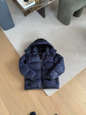 Mörkblå dunjacka från Polo Ralph Lauren - Snygg mörkblå dunjacka från Polo Ralph Lauren med röd logga på bröstet. Jackan har huva, dragkedja framtill, bröstficka med dragkedja och två sidofickor. Perfekt för kalla vinterdagar och riktigt skön att bära.och självklart äkta,tveka inte på att höra av er ❤️Jag är 179 cm och ni ser på bilden hur den sitter på mig som är 179 😁men strl S,priset går självklart att diskutera