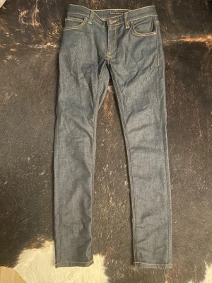 Mörkblå jeans från Nudie - Säljer ett par mörkblå jeans från Nudie Jeans med slim passform och klassisk fem fick design. Jeansen har kontrastsömmar i gult och är tillverkade i rå denim för en clean look. Perfekta för dig som gillar stilrena och tidlösa jeans. Använd ca 5 ggr☺️🙌 skriv gärna vid frågor!