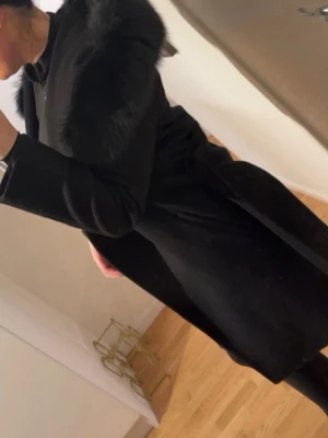 Svart kappa från Hollies - Stilren svart kappa med stor luva kantad av mjuk fuskpäls. Kappan har knytskärp i midjan och raka ärmar. Perfekt för kalla dagar när du vill hålla dig varm och se snygg ut. Klassisk och tidlös design som passar till många olika outfits.