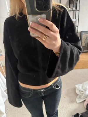Svart croppad tröja med vida ärmar - Säljer en svart croppad tröja med vida ärmar och ribbad nederkant. Tröjan har en enkel och stilren design som passar perfekt till jeans eller kjol. Materialet ser mjukt och bekvämt ut, perfekt för en avslappnad look.