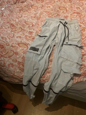 Ljusgrå cargobyxor Approved - Säljer ett par ljusgrå cargobyxor från Approved med streetwear-vibe. Byxorna har flera stora fickor, svarta detaljer och dragsko i midjan. Materialet känns mjukt och bekvämt, perfekta för en avslappnad stil. Snygga detaljer med patch och metallring.