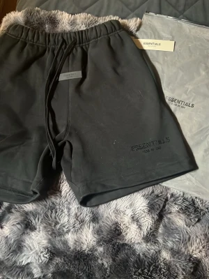 Svarta shorts från Essentials Fear of God - Svarta shorts från Essentials Fear of God med diskret logga på benet och snörning i midjan. Klassisk loose fit och tillverkade i mjukt bomullsmaterial. Perfekta för chill dagar eller streetwear-stil.