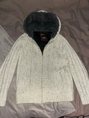Vit/beige stickad cardigan - Mysig vit stickad jacka från Petroleum med kabelstickat mönster och dragkedja framtill. Jackan har en stor huva fodrad med mjuk grå fuskpäls och svart innerfoder. Perfekt för kyliga dagar och riktigt skön att bära.