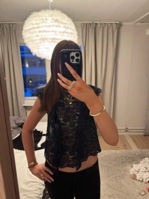 Svart spetsig croppad topp - Säljer en svart croppad topp i spets med ärmlös design och fina volangdetaljer längs axlarna. Toppen är transparent och har ett elegant mönster i spets som ger en trendig look. 