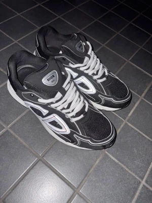 Svart vita dior B30 - Snygga svarta Dior B30 sneakers med vita och silverfärgade detaljer samt vita skosnören. Skorna har en sportig design med mesh och syntetmaterial, samt tydlig Dior-logga på plösen. Perfekta för dig som vill ha en exklusiv och trendig look.