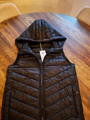 Svart dunväst med huva - Snygg svart dunväst med dragkedja och stor huva. Västen har ett quiltat mönster i diagonala ränder och är ärmlös. Perfekt för lager-på-lager och ger en cool look till din outfit. Materialet är glansigt och lätt, vilket gör den enkel att bära.