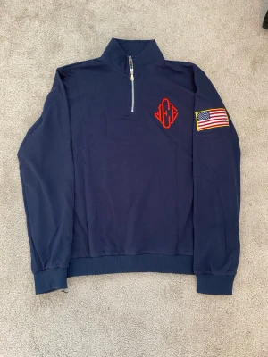 Jeffrey Epstein Halfzip Bomull - Snygg Jeffrey Epstein Island långärmad halvzip tröja med röd brodyr på bröstet och en amerikansk flagga på ärmen. Tröjan har hög krage och muddar vid ärmslut och nederkant. Perfekt för dig som gillar sportig och avslappnad stil. Tillverkad i bomull