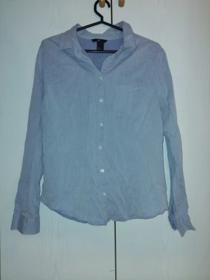 Ljusblå skjorta från H&M - Klassisk ljusblå skjorta från H&M med lång ärm och knappar framtill. Skjortan har en avslappnad passform och är tillverkad i ett mjukt bomullstyg. Perfekt att styla till jeans eller kjol för en clean look.