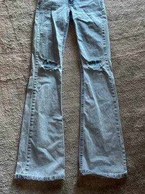 Ljusblå bootcut jeans med slitningar - Säljer ett par ljusblå bootcut jeans med slitna hål på knäna och rosa broderade detaljer på bakfickorna från Nelly. Jeansen har klassisk femficksmodell och är tillverkade i denim. Perfekta för en avslappnad och trendig look.