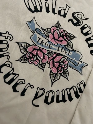 Vit tröja  med tryck och rosor - Vit tröja  med stort tryck i svart, blått och rosa. Motivet har texten 'Wild Soul Forever Young True Love' och dekoreras av rosa rosor och ett blått band. Aldrig använd alltså helt ny med prislappen kvar🩷