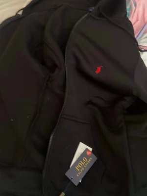 Svart hoodie från Polo Ralph Lauren - Snygg svart hoodie från Polo Ralph Lauren med dragkedja och huva. Klassisk röd logga broderad på bröstet och fickor framtill. Tillverkad i mjukt material som passar perfekt till en avslappnad stil.