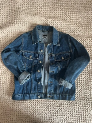 Klassisk blå jeansjacka - Snygg blå jeansjacka från VRS med raka linjer och två bröstfickor med knappar. Jackan har klassisk krage och silverfärgade knappar. Perfekt att slänga över en hoodie eller t-shirt.