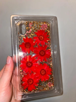 iPhone XS Max mobilskal - Unikt mobilskal med stora röda blommor och inslag av guldglitter. Skalets bakgrund är transparent vilket gör att blommorna och glittret verkligen poppar. Passar perfekt för dig som vill ha ett färgstarkt och personligt mobilskal. Materialet är troligtvis plast eller TPU.