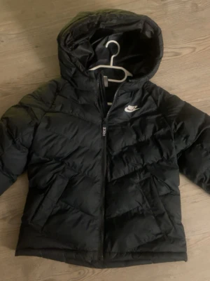 Svart jacka från Nike - Snygg svart pufferacka från Nike med huva och dragkedja framtill. Jackan har ett stilrent Nike-logga på bröstet. Perfekt för kalla dagar och enkel att matcha med olika outfits. 
