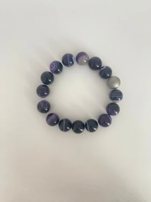 Lila pärlarmband med silverdetalj - Snyggt armband med runda pärlor i olika nyanser av lila och vitt, samt en unik matt silverfärgad pärla som accent. Pärlorna har ett coolt mönster med ränder och är trädda på elastisk tråd för enkel användning. Perfekt för dig som gillar färg och vill sticka ut.