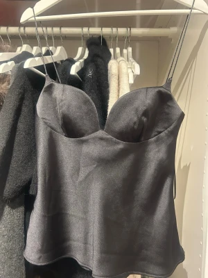 Svart satinlinnetopp från Zara - Snygg svart linnetopp från Zara i glansigt satinmaterial. Toppen har tunna axelband och formade kupor som ger en feminin siluett. Perfekt att styla med jeans eller kjol för en trendig look.