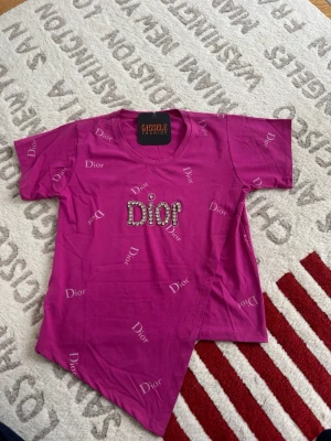 Rosa Dior t-shirt med strassdetaljer - Säljer en rosa t-shirt från Giselle Fashion med Dior-tryck över hela och stora strassdetaljer på bröstet. T-shirten har korta ärmar, rund halsringning och en unik asymmetrisk nederkant. Perfekt för dig som vill sticka ut med en trendig och färgstark look.