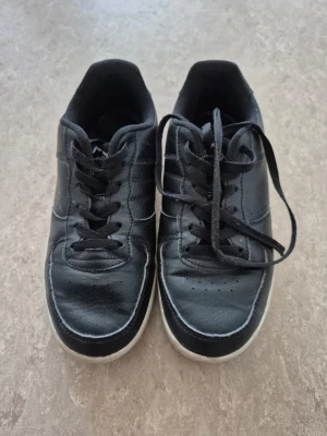 Sneakers - Klassiska svarta sneakers från Nonation med vita sulor och svarta skosnören. Skorna har en låg profil och är tillverkade i syntetmaterial med diskreta sömdetaljer. Perfekta för en avslappnad och stilren look.