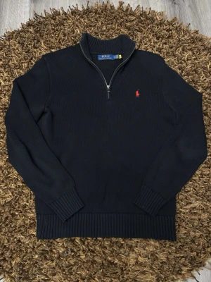 Svart tröja Polo Ralph Lauren - Svart quarter zip tröja från Polo Ralph Lauren med halv dragkedja och röd broderad logga på bröstet. Tröjan har ribbade muddar och hög krage, perfekt för lager på lager. Sydd vid vänstra ärmen.