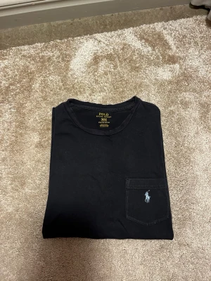 Mörkblå t-shirt från Polo Ralph Lauren - Stilren mörkblå t-shirt från Polo Ralph Lauren med rund halsringning och bröstficka. T-shirten har en broderad logga i ljusblått på fickan och är gjord i stretchig bomull för extra komfort. Hör av er vid frågor eller funderingar! 