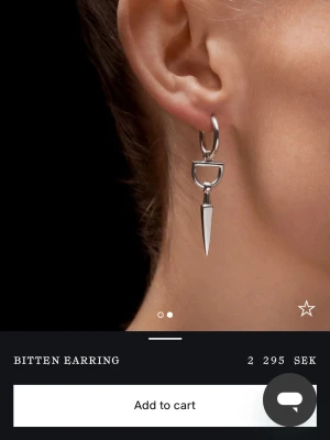Bitten Earring i silver - Säljer ett stilrent örhänge från maria Nilsdotter. Örhänget har en unik design med en rund ring och en spetsig, avlång hängande detalj. Perfekt för dig som gillar minimalistiska och edgy smycken. Säljer mitt andra favorit örhänge nu, kommer inte till användning längre.