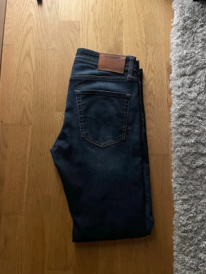 Mörkblå jeans från Jack & Jones - Snygga mörkblå jeans från Jack & Jones med klassisk femficksdesign och bruna, W28 L32, kontrastsömmar. Jeansen har en läderpatch med logga bak i midjan och är tillverkade i ett mjukt denimtyg som passar perfekt till vardagslooken. Modellen är Slim Straight/Tim.