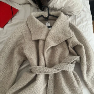 Beige teddy-kappa från Gina Tricot - Mysig beige teddy-kappa från Gina Tricot med bred krage och knytskärp i midjan. Kappan har en fluffig, texturerad yta och en avslappnad passform som ger en trendig look. Perfekt för kalla dagar när du vill vara både varm och snygg.ny pris 600
