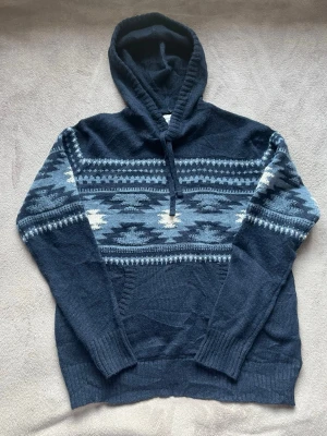 Mörkblå stickad hoodie med mönster - Mysig mörkblå stickad hoodie med huva och snyggt geometriskt mönster i ljusblått, vitt och grått över bröstet och armarna. Tröjan har långa ärmar, ribbade muddar och en klassisk känguruficka framtill. Perfekt för kalla dagar och chill stunder.