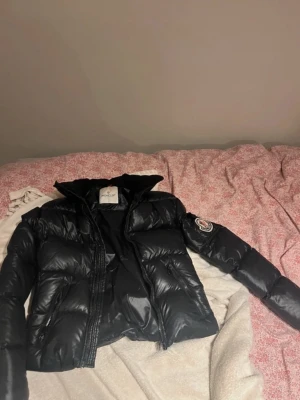 Svart dunjacka. Liknar Moncler! - Snygg svart dunjacka Moncler dupe med glansig finish och klassisk Moncler-logga på ärmen. Perfekt för kalla vinterdagar och riktigt varm tack vare dunfyllningen. Ej äkta!!! I bra skick. Finns lite defekter som man ser på bilden 