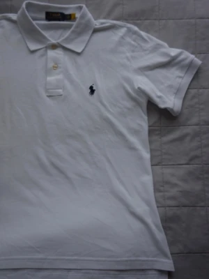 Pike | Ralph Lauren - Klassisk vit pikétröja från Ralph Lauren med broderad logga på bröstet. Tröjan har krage, knappar framtill och korta ärmar. Perfekt till jeans eller shorts för en clean och stilren look. Tröjan är i Ralph laurens klassiska custom slim fit! 