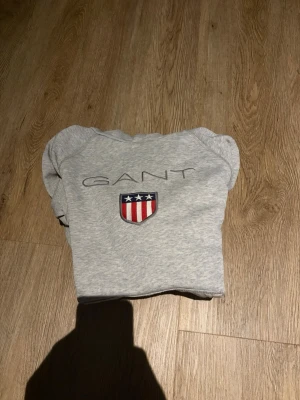 Grå hoodie från GANT med logga - Säljer en stilren grå hoodie från GANT med deras klassiska broderade logga och sköld i rött, vitt och blått på bröstet. Hoodien har huva och ribbade muddar vid ärmslut och nederkant. Perfekt för en avslappnad och snygg look.