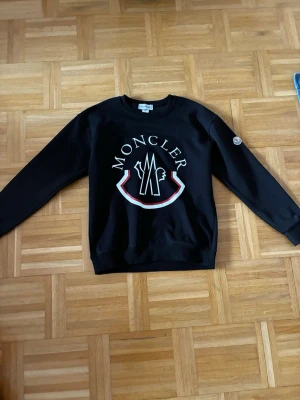 Svart sweatshirt från Moncler - Säljer en svart sweatshirt från Moncler med stort vitt och rött logotryck på bröstet och rund hals. Tröjan har ribbade muddar vid ärmslut och nederkant samt en patch på ena ärmen. Perfekt för dig som gillar streetwear och exklusiva märken.
