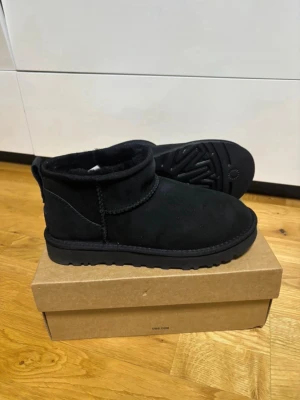 Svarta UGG mocka - Säljer ett par svarta UGG i mjuk mocka med fårskinnsfoder. Klassisk låg modell med grov sula och diskret UGG-logga bak på hälen. Perfekta för att hålla fötterna varma och sköna under kalla dagar.