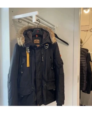 Vinterjacka från Parajumpers stl S - Snygg svart parkas från Parajumpers med avtagbar huva och fuskpäls, flera praktiska fickor och ikonisk gul rem vid kragen. Jackan har dragkedja och knappar framtill samt detaljer i metall. Perfekt för kalla dagar och riktigt street.