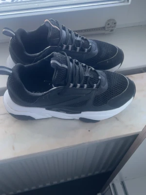 Dior b22 - Snygga svarta Dior sneakers med vit sula och meshdetaljer för extra ventilation. Skorna har svarta och gråa snören samt en chunky sula som ger en modern look. Perfekta för dig som vill ha både stil och komfort i vardagen.