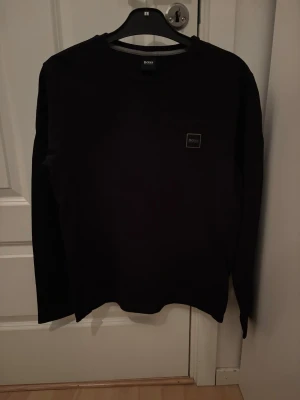 Svart sweatshirt från BOSS - Snygg svart sweatshirt från BOSS med rund halsringning och diskret logotyp-patch på bröstet. Tröjan har en klassisk passform och långa ärmar, perfekt för en stilren och enkel look.