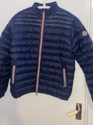 Mörkblå pufferjacka från Moncler - Snygg mörkblå pufferjacka från Moncler med quiltad design och raka linjer. Jackan har dragkedja och fickor med röd, vit och blå randiga detaljer samt Moncler-logga på ärmen. Perfekt för kalla dagar och riktigt trendig look.