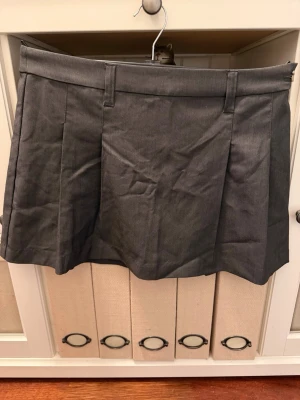 Mörkgrå kortkjol med veck - Snygg mörkgrå kortkjol med bred linning och markerade veck framtill och baktill. Kjolen har hällor för bälte och en stilren, minimalistisk look som passar till många olika outfits.