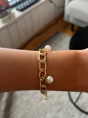 Guldarmband med pärlor - Snyggt armband i guldfärgad metall med stora länkar och hängande vita pärlor. Perfekt accessoar för att lyfta din outfit med en trendig och elegant touch. Armbandet har en chunky kedjedesign och fyra pärlor som ger en cool kontrast.