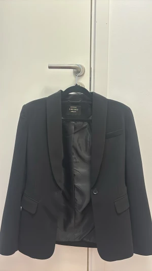 Svart kavaj från New Yorker - Stilren svart kavaj från Amisu med klassisk sjalkrage och en knapp framtill. Kavajen har två fickor med lock och en bröstficka. Fodrad insida i blankt tyg ger en snygg finish. Perfekt för en dressad look.