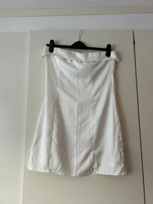 Vit kort klänning med slits och uppvikt kant - Säljer en vit klänning med uppvikt kant upptill och en liten slits framtill. Klänningen har en A-linjeformad siluett och är enkel men stilren. Perfekt för dig som gillar minimalistisk stil.