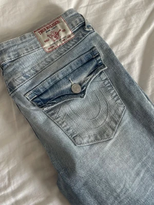 True Religion ljusblå jeans - Ljusblå jeans från True Religion med klassisk femficksdesign och ikoniska kontrastsömmar på bakfickorna. Jeansen har raka ben och är gjorda i mjuk denim. Snygg detalj med knappstängning på fickan och tydlig logopatch bak i midjan.